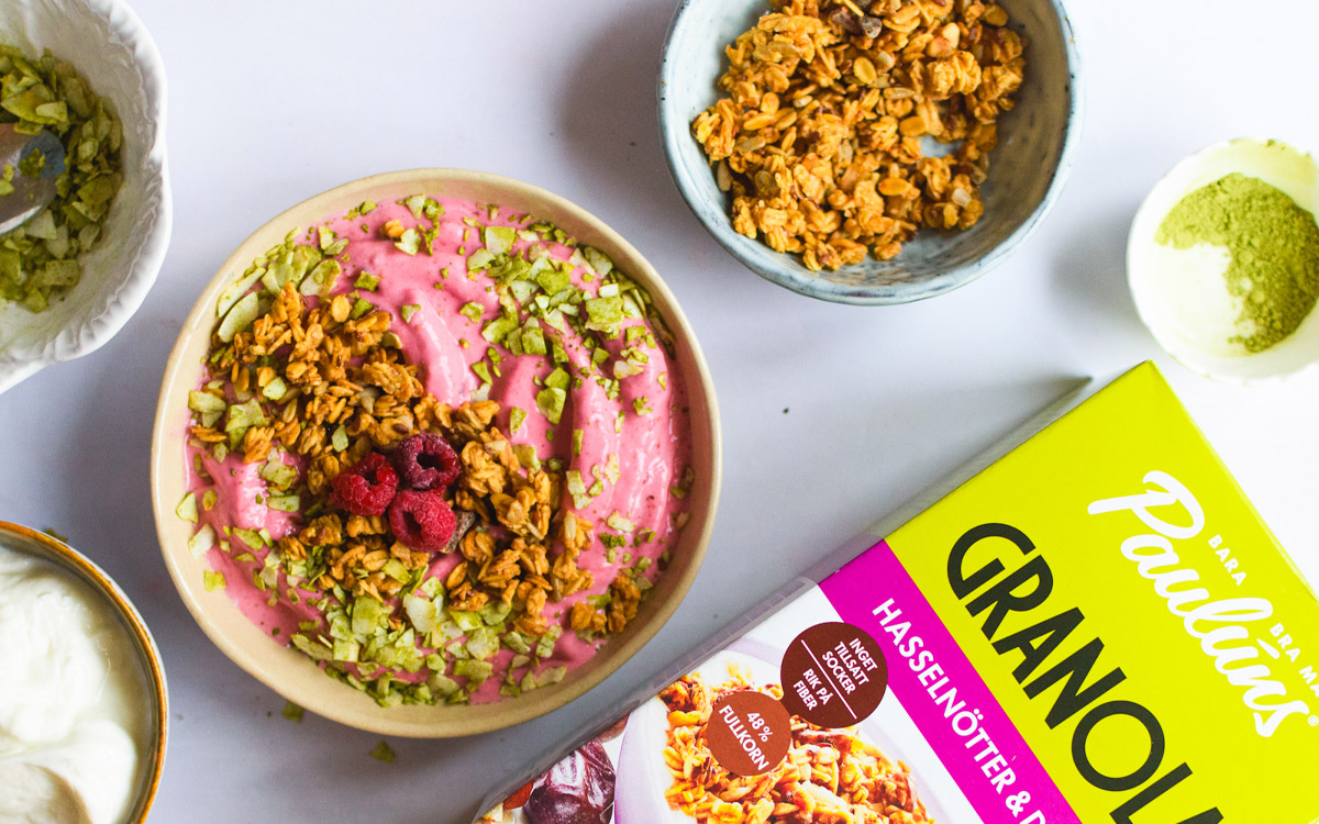 Skål med frozen yoghurt toppad med Pauluns Granola Hasselnötter & Dadlar, matchapulver och hallon.